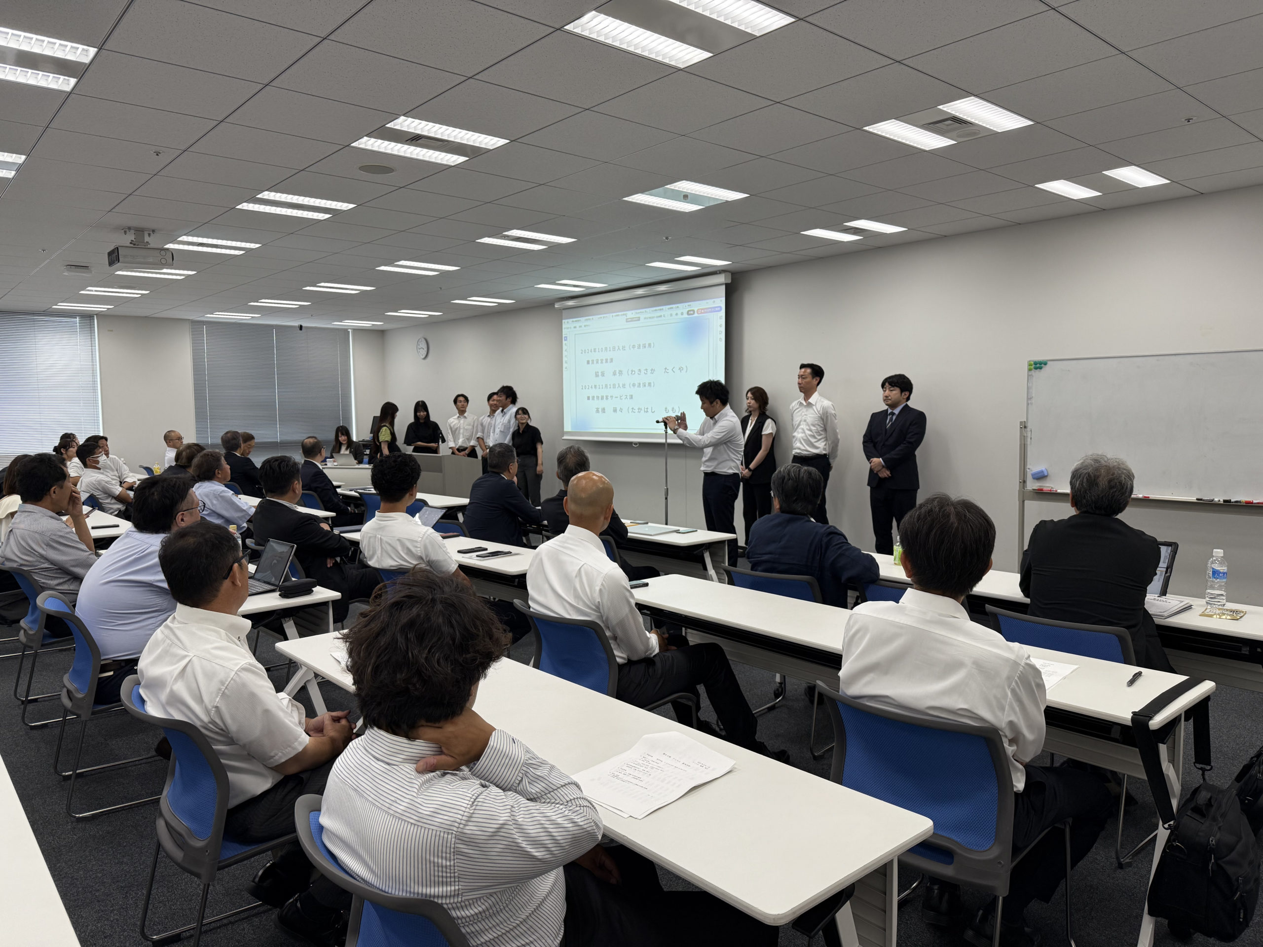 2025年9月に行われたアミックス「第69期経営方針発表会」の画像の2枚目。各部署の代表者が登壇し、一人が発表しており、数人は待機している。後ろ姿が見える社員は発表者に注目して話を聞いている。