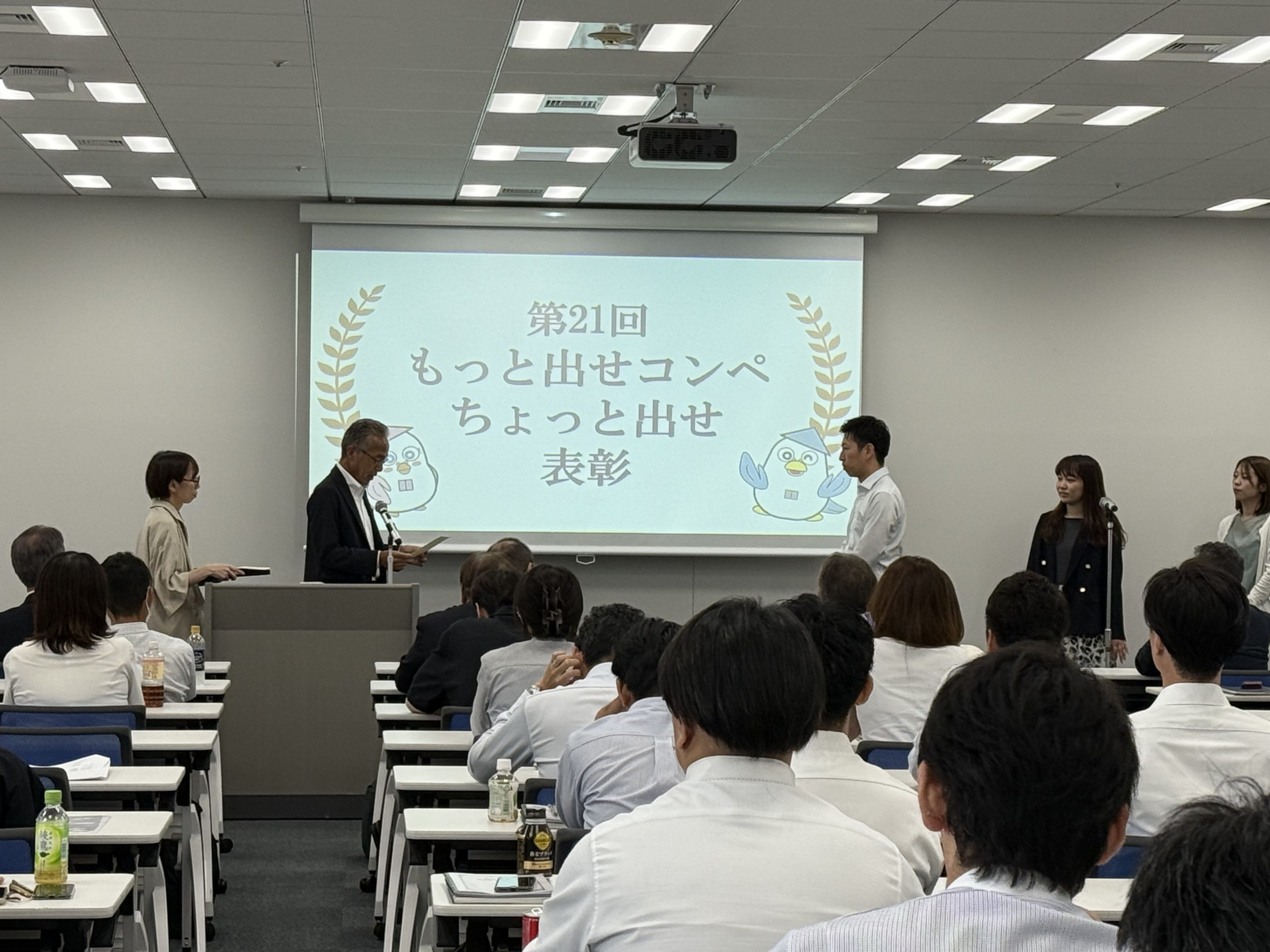 2025年9月に行われたアミックス「第69期経営方針発表会」の画像の3枚目。正面のスクリーンには「第21回もっと出せコンペ・ちょっと出せ 表彰」の文字が映されている。スクリーン左手にはプレゼンターのアミックス中町役員と賞状をもって待機している社員がおり、右手には受賞者の男性社員が表彰を受けている。その後ろには2名の女性社員がならんでおり、表彰の順番を待っている。後ろ姿が見える社員はその様子に注目している。