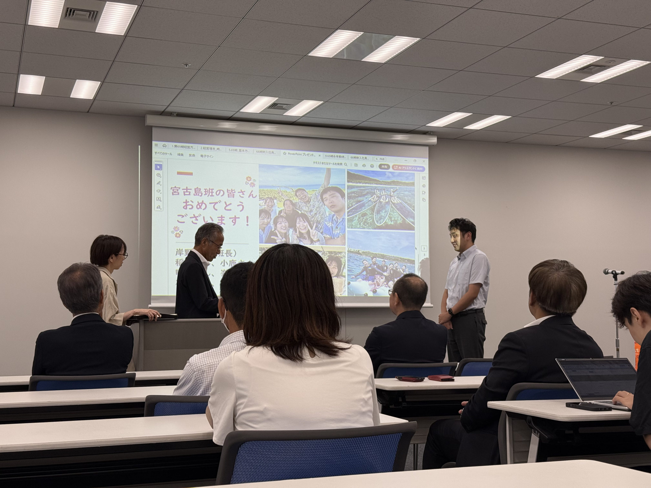 2025年9月に行われたアミックス「第69期経営方針発表会」の画像の3枚目。正面のスクリーンには「宮古島の皆さんおめでとうございます！」の文字が映されている。スクリーン左手にはプレゼンターのアミックス中町役員と賞状をもって待機している社員がおり、右手には受賞者の男性社員が表彰を受けている。後ろ姿が見える社員はその様子に注目している。